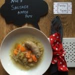 Photo of the Nic's Pressure Cooker Sausage Apple Pot seen here served on a deep white plat with a grey napkin wrapped in a red and white polka dot ribbon to the right and the Feisty Tapas logo on a card on the top right. There's also a chalkboard with the name of the recipe written on it