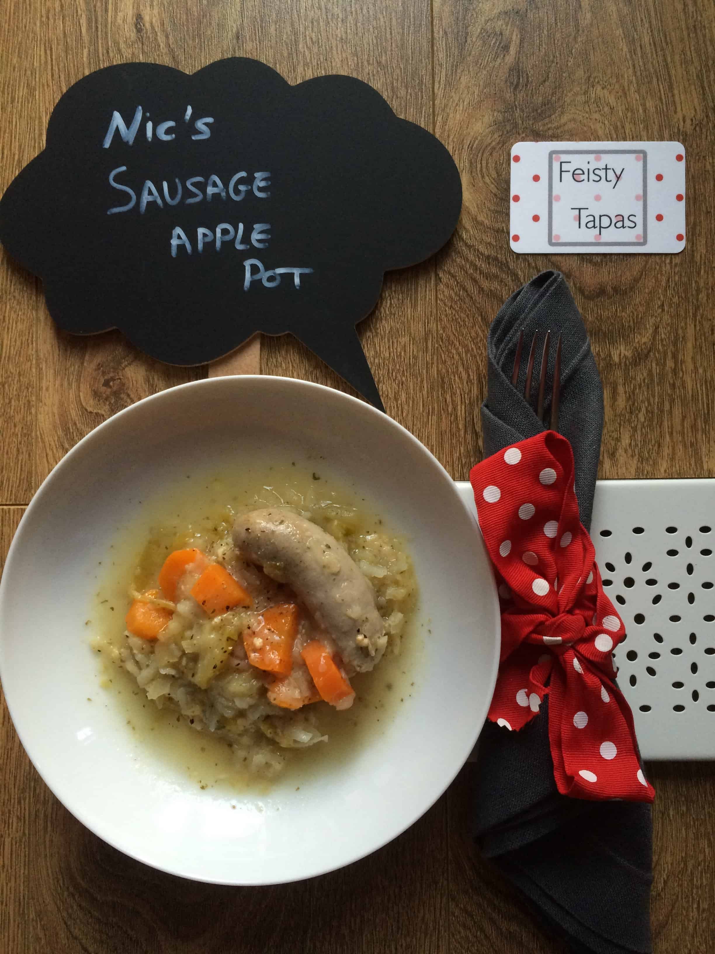 Nic's Instant Pot Pressure Cooker Sausage Apple Pot. A delicious dish seen here served on a deep white plat with a grey napkin wrapped in a red and white polka dot ribbon to the right and the Feisty Tapas logo on a card on the top right. There's also a chalkboard with the name of the recipe written on it