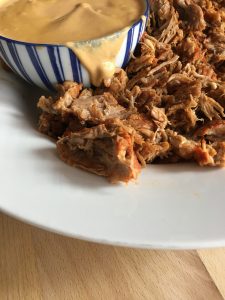Instant Pot Pulled Pork with Creamy Paprika Sauce by Feisty Tapas. The pulled pork is served in a large white round serving dish and the sauce is looking delicious oozing from the side of a smaller blue and white striped bowl. On a wooden table