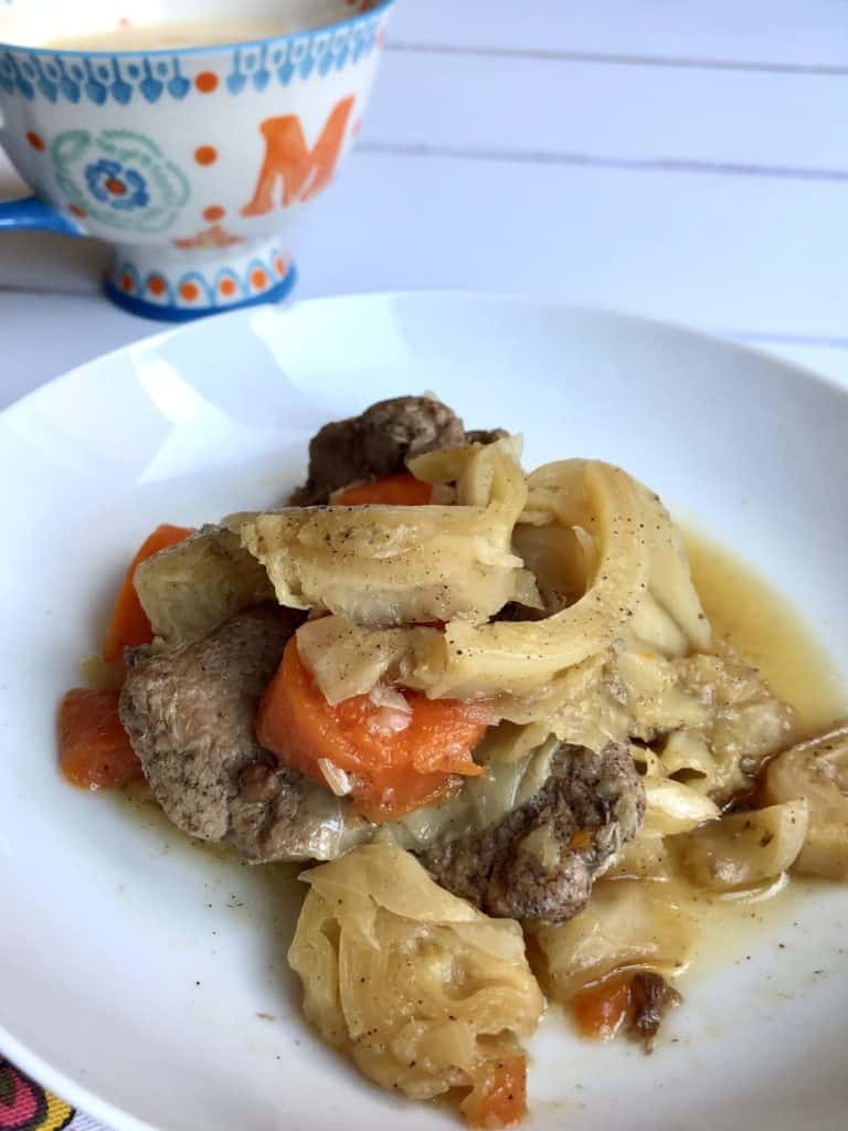 Instant Pot Pork and Cabbage recipe by Feisty Tapas - served on a white plate with a tea towel with orange pom poms and a mug with the initial M in the background