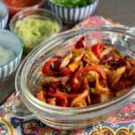 Air Fryer Smoky Chicken Fajitas in an oval pyrex dish with handles, on a colourful tea towel with the condiments behind it