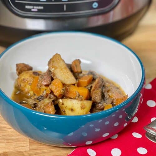 Pressure Cooker Vegan Mushroom Stew seen inside a bowl, blue on the outside and white on the inside. There is a cloth napkin with white polka dots on red background on the bottom right. The bowl is on a wooden surface with a Ninja Foodi Max 9 in 1 in the background