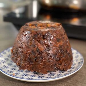 Finished Christmas Pudding turned onto a white and blue patterned plate on a brown surface with pressure cookers in the background