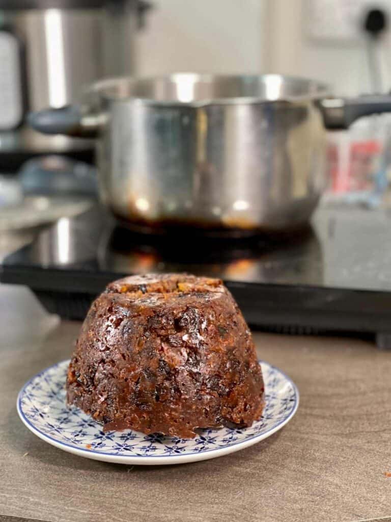 A pressure cooked Christmas Pudding show on a white and blue patterned plate on a brown surface with a standalone induction hob in the background with the pot of a pressure cooker on top