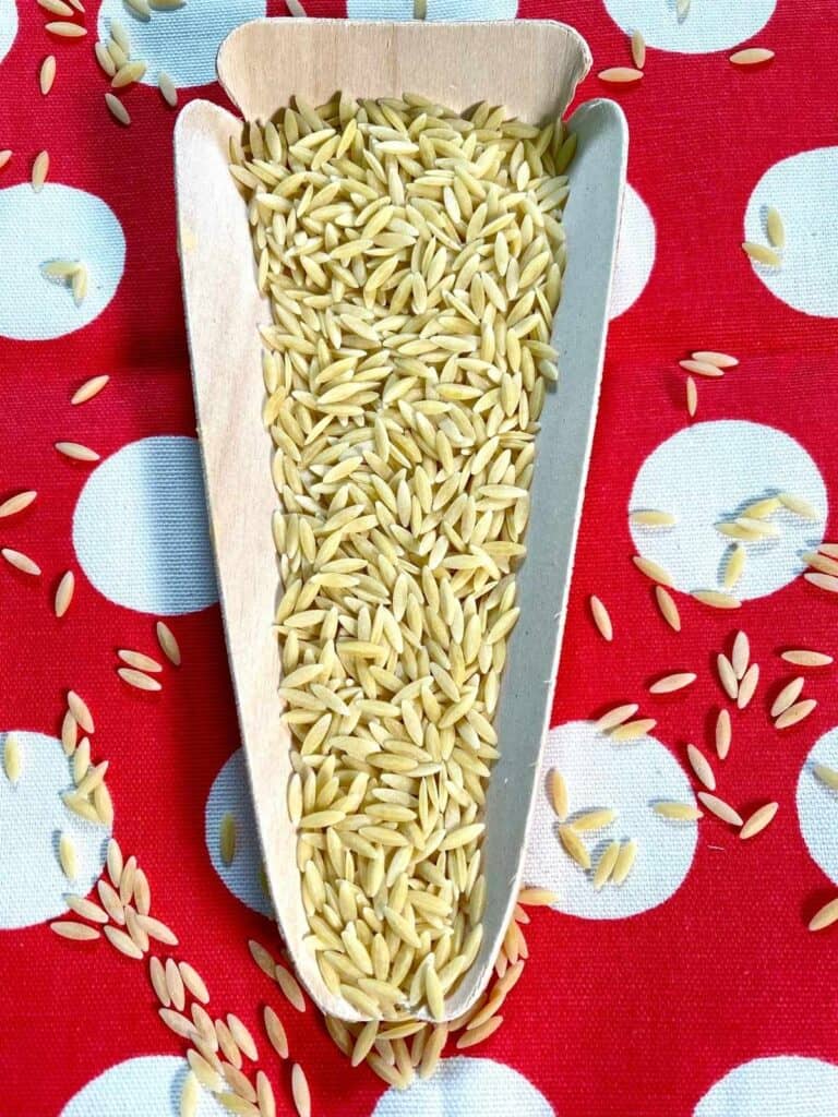Orzo pasta seen from above on a wooden triangle against a red and white polka dot background, with teardrop orzo pasta overflowing