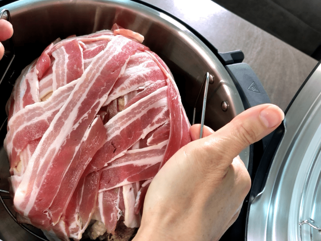 Turkey crown covered in bacon being lowered into inner pot to air fry. Seen from above with hands lowering it