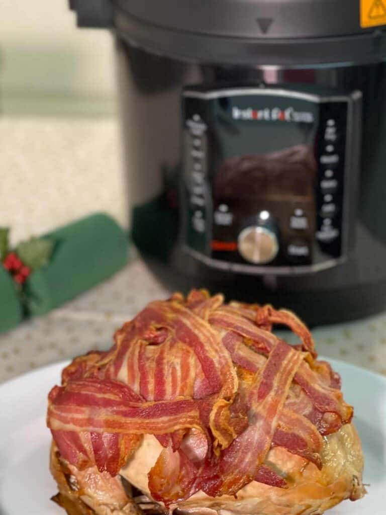 Pressure cooked Instant Pot Turkey Crown ready to carve, still covered in bacon, seen on a white dish with the Instant Pot Pro Crisp in the background and a green christmas cracker