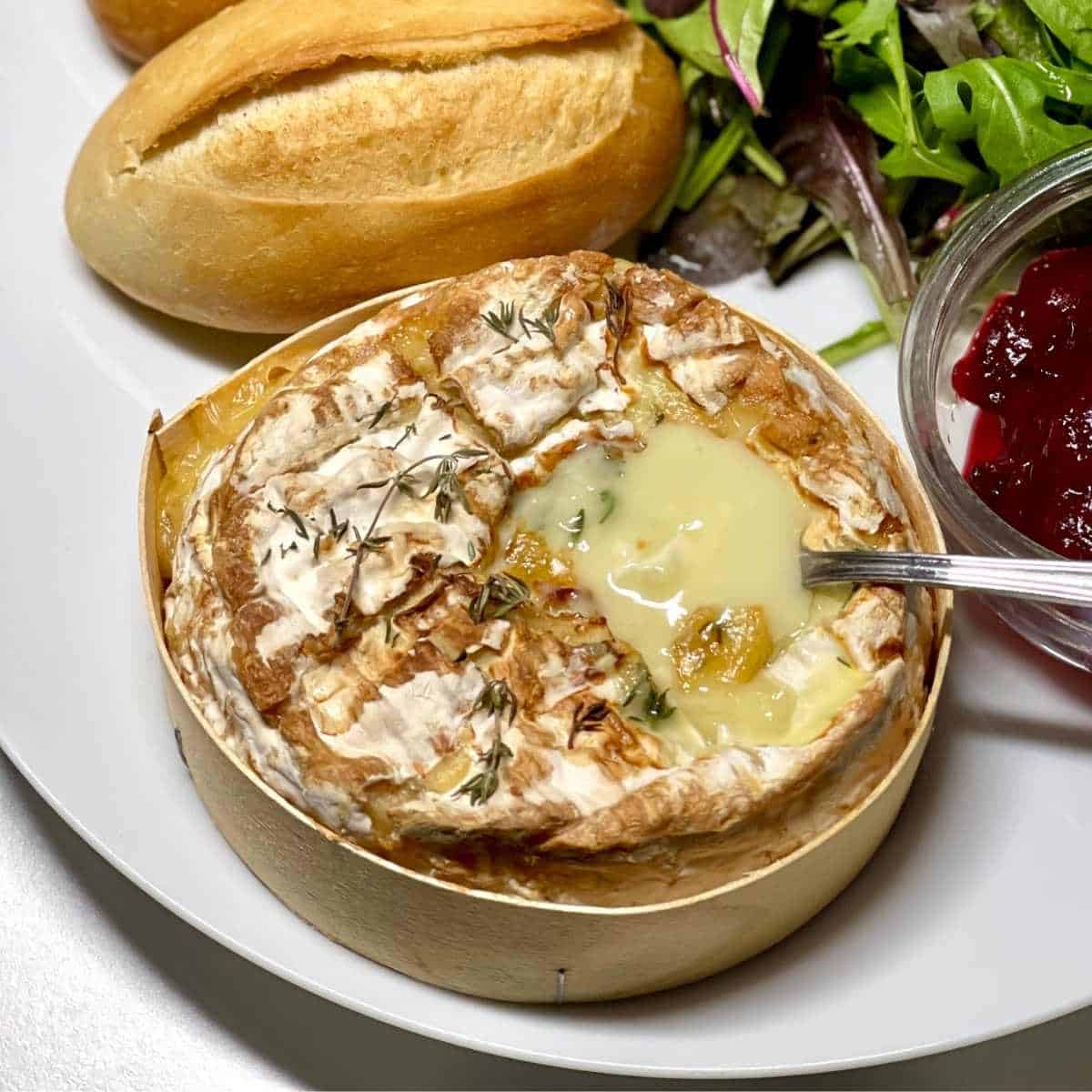Air Fryer Baked Camembert in its wooden box, oozing with a stainless steel spoon sticking on the right side, all on a white plate with a crusty roll and some cranberry sauce