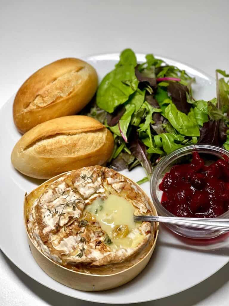 Baked camembert still in the wooden box seen from above on a white plate with a little bowl with cranberry sauce salad and two crusty rolls