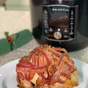Pressure Cooked Turkey Crown covered in crispy bacon that has been air fried with the air fryer lid. On a white oval dish with a spotty beige tablecloth with gold polka dots and a green Christmas cracker. in the background there is an Instant Pot Pro Crisp