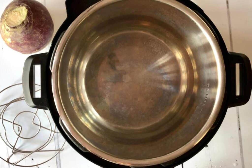 Instant Pot Duo seen from above, with a whole uncooked swede on the top left and the trivet or steamer rack on the bottom left. Against a white background