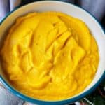 Vibrant orange pressure cooker carrot and mash in a white bowl with a dark blue rim, a hand in a grey sweater can be seen holding it