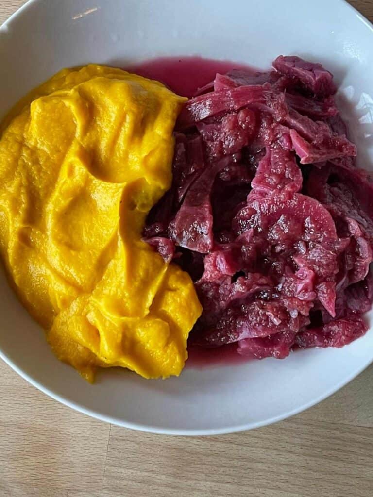 Carrot and Swede Mash served on a white plate next to red cabbage, against a wooden surface