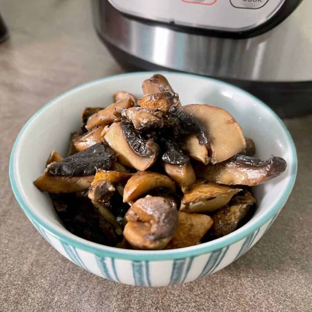 Pressure Cooker Garlic Mushrooms (Spanish Setas al Ajillo) seen in a white and blue/green stripy bowl on a brown surface with an Instant Pot Duo in the background