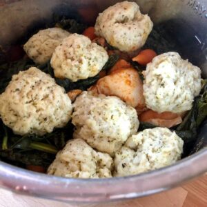 The pressure cooked chicken and vegetables topped with dumplings is seen from above still inside the stainless steel inner pot of the Instant Pot, on a light wooden surface