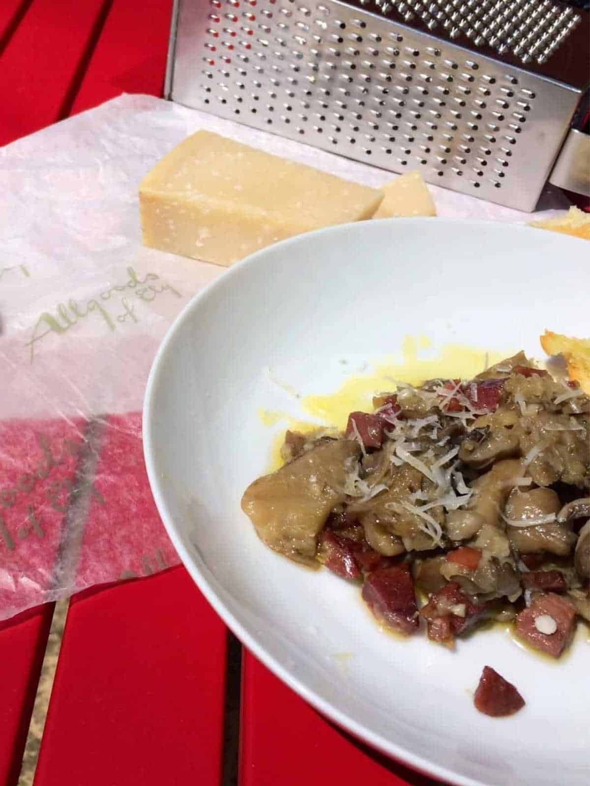 Thermomix Spanish Mushrooms with serrano ham, setasl al ajillo, served on a white plate against a red slatted surface. There is a metallic cheese grater at the top of the photo and a wedge of parmesan cheese resting on wax paper