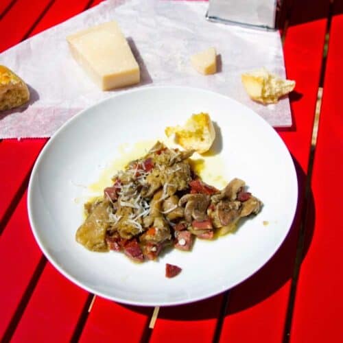 Thermomix Garlic and Serrano Ham Mushrooms (Setas al ajillo) seen on a white plate placed on a red slatted table with a wedge on parmesan in the background and some crusty bread