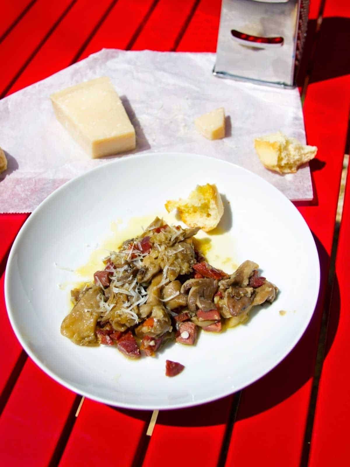 Thermomix Spanish Mushrooms with serrano ham, setasl al ajillo, served on a white plate against a red slatted surface. There is a metallic cheese grater at the top of the photo and a wedge of parmesan cheese resting on wax paper with some crusty bread