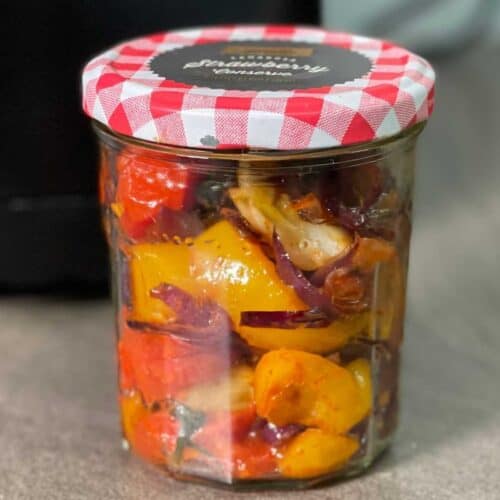 Air Fryer Roasted Vegetables stored in a glass jam jar with red and white gingham lid on a brown surface with a black air fryer in the background