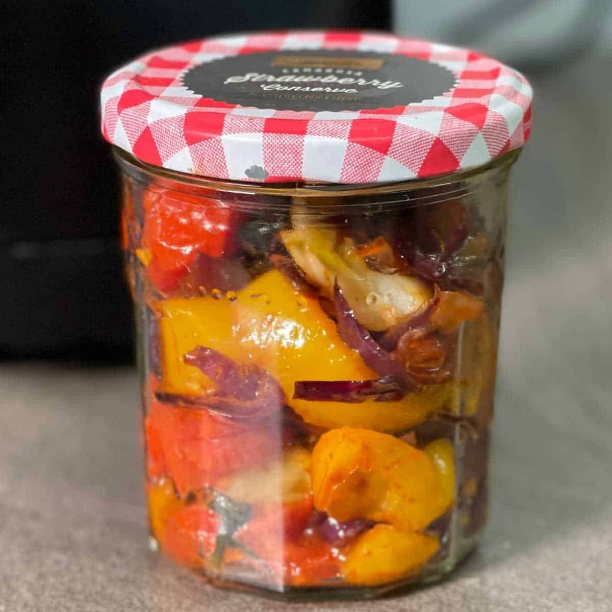 Air Fryer Roasted Vegetables stored in a glass jam jar with red and white gingham lid on a brown surface with a black air fryer in the background