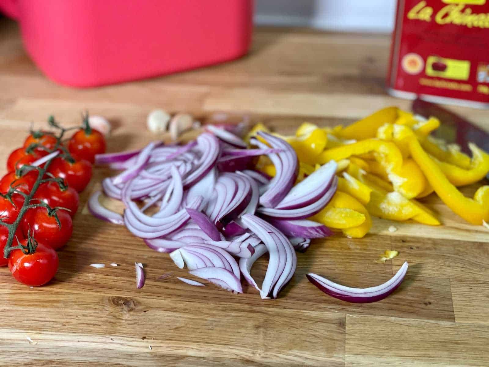 Ingredients for Air Fryer Roasted Vegetables laid out on a light coloured wooden chopping board: cherry tomatoes on the vine, sliced pepper and red onion, whole unpeeled garlic cloves
