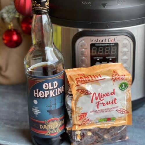 Bottle of dark rum from Aldi and bag of dried fruit mix from Aldi too with an Instant Pot Duo and a Christmas tree with red baubles in the background