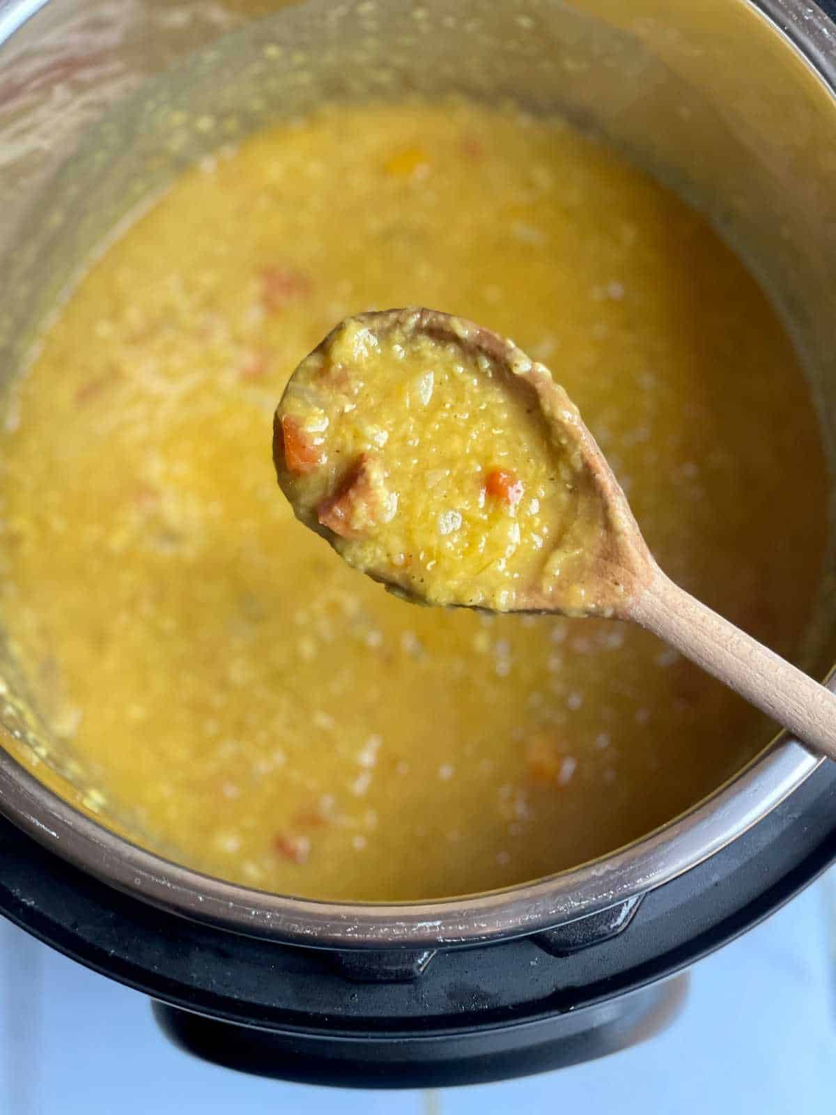 A wooden spoon is seen from above holding some of the red lentil and chorizo soup, underneath is an Instant pot electric pressure cooker
