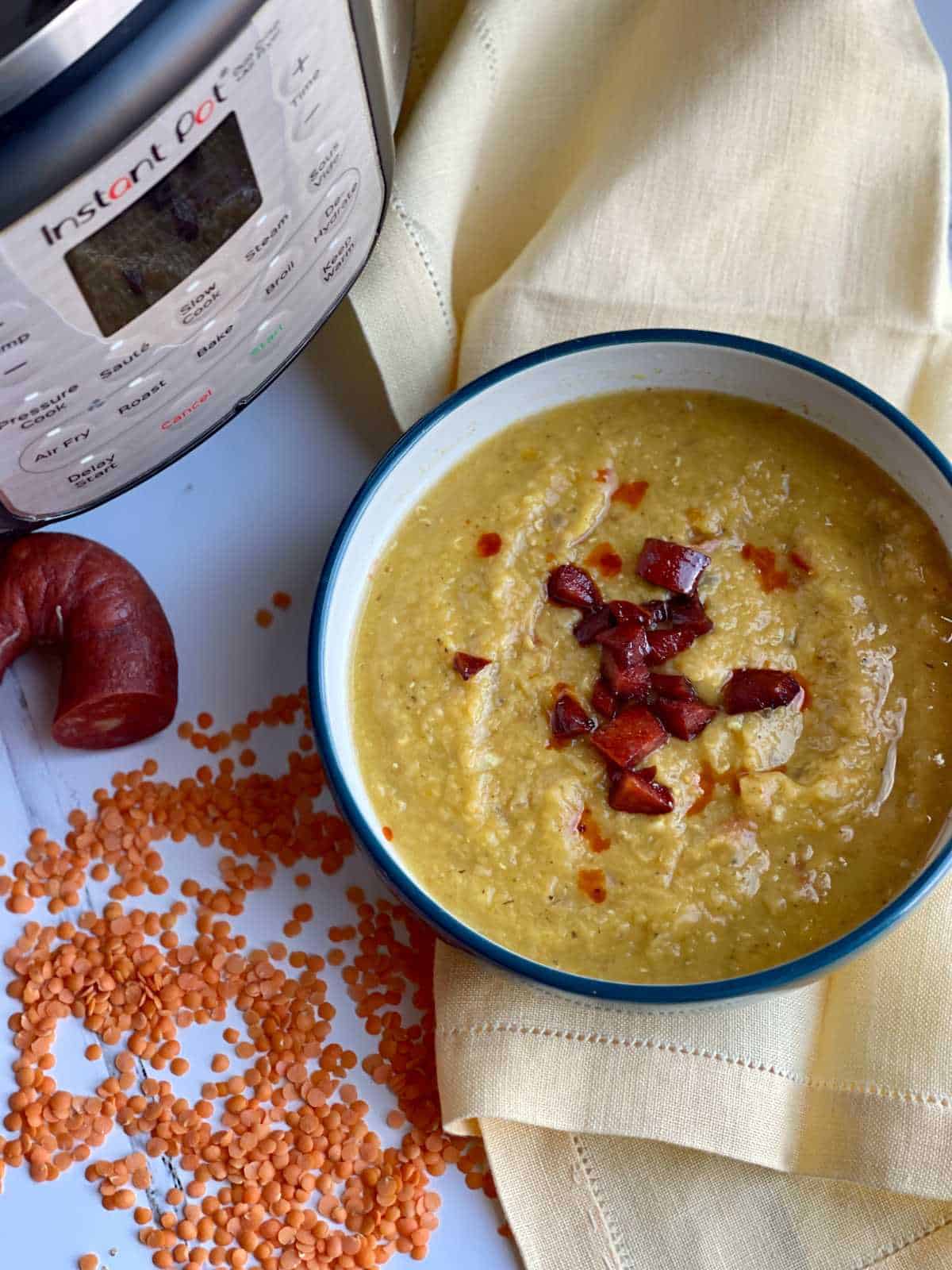 Pressure Cooker Chorizo and Red Lentil Soup seen from above inside a blue bowl with white inside. The bowl is resting on a yellow linen napkin. There are red lentils scattered on the white surface on the left, a chorizo sausage hoop with a chunk missing and an Instant Pot Duo Crisp