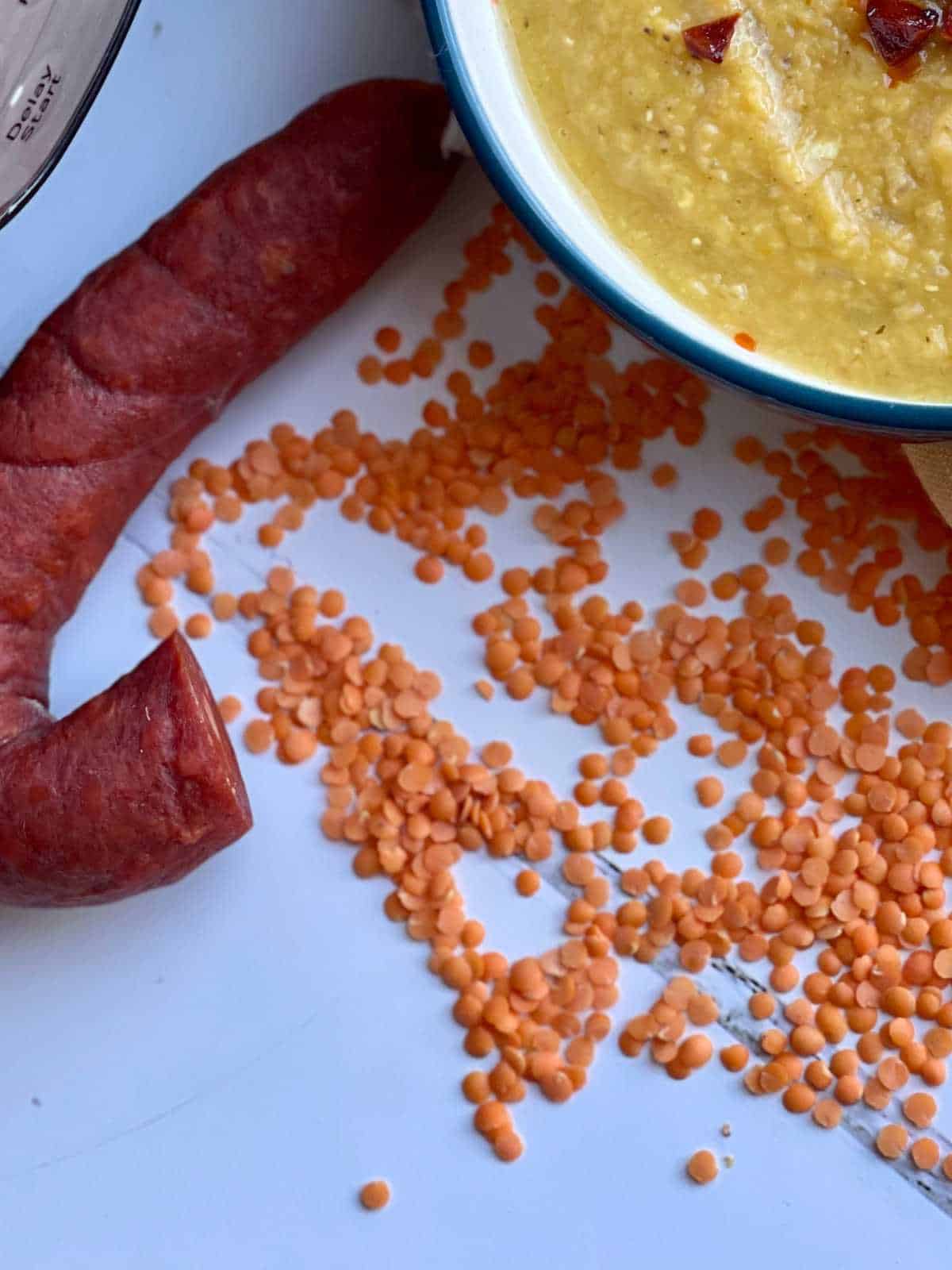 red lentils scattered on a white surface with a chorizo loop on the top left and a bowl of soup on the top right, all seen from above