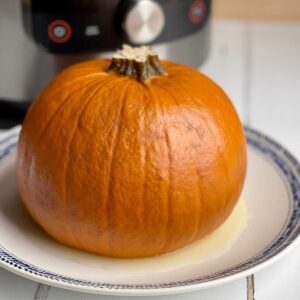 Photo of a whole pressure cooked pumpkin sitting on a white plate in front of a Ninja Foodi 15 in 1 electric pressure cooker