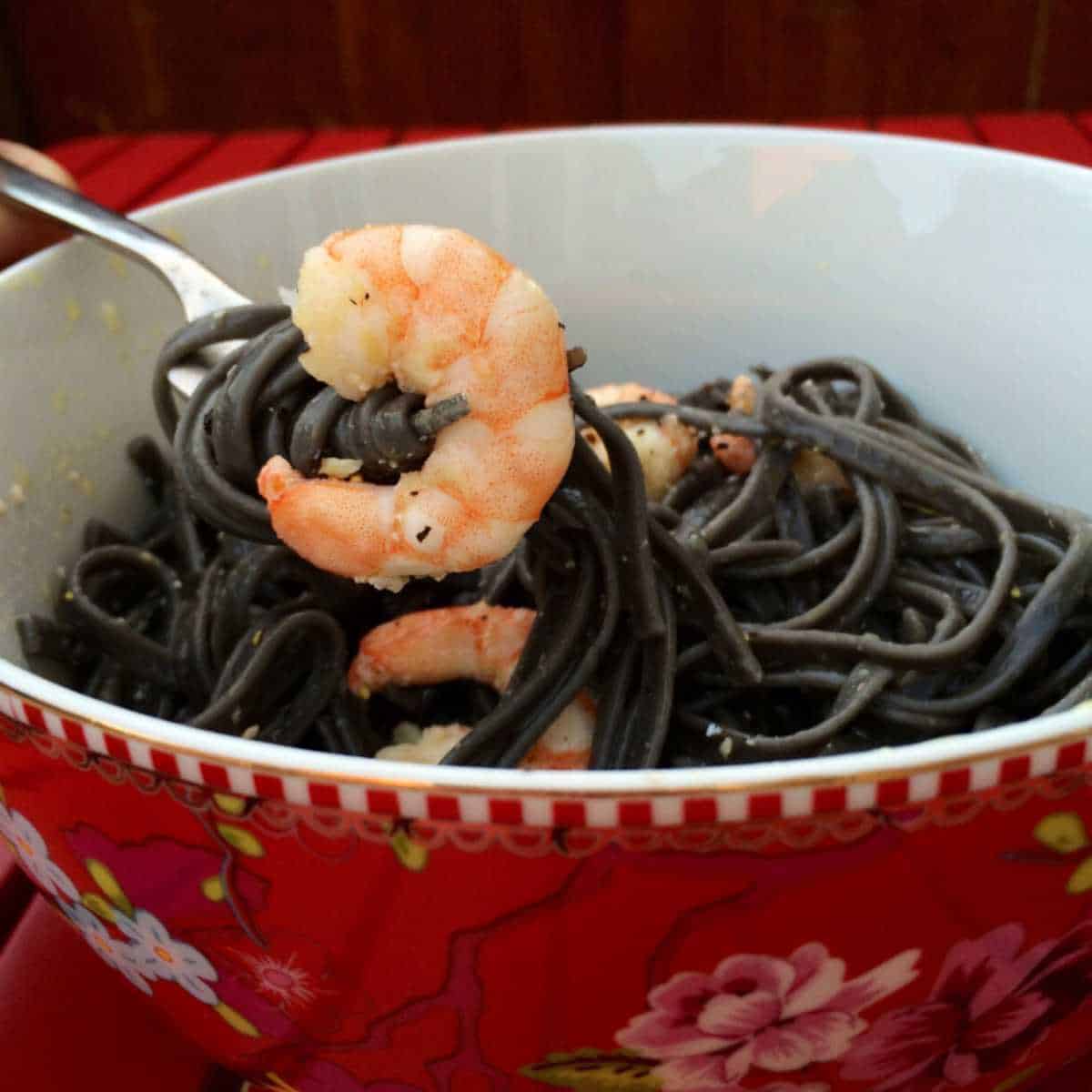 Photo of a large prawn being held up by a fork with black squid ink pasta twisted around it, held above a pretty flowery Pip Studio bowl