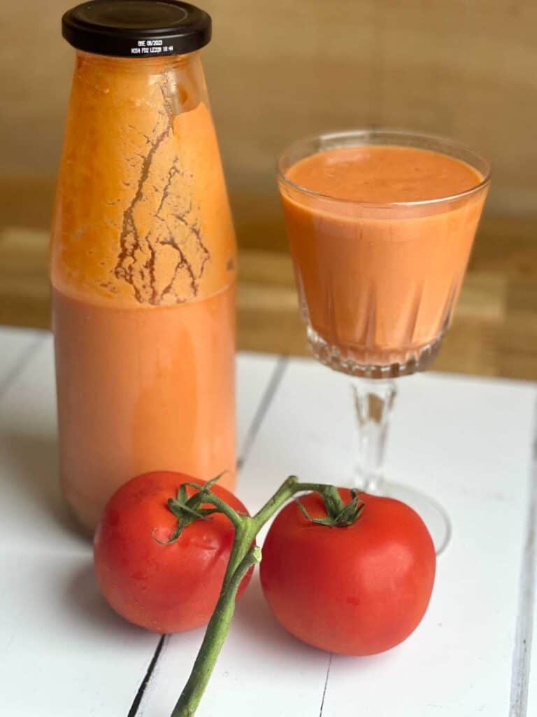 On the right there is a pretty stem glass full of gazpacho, on the left there is an old passata bottle half full with gazpacho and in front of them there are two ripe tomatoes on the vine