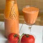 Photo of Authentic Spanish Gazpacho blended in the Thermomix and served in a pretty glass shown with a couple of red tomatoes on the vine in front of it and a bottle half full of gazpacho ready to store in the fridge