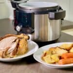 Photo of the whole chicken and the vegetables served in two large plates with the Ninja 14 in 1 in the background. The vegetables include potatoes, carrots and parsnips. The chicken looks juicy
