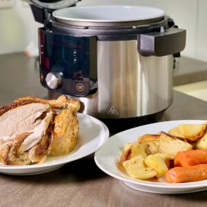 Photo of the whole chicken and the vegetables served in two large plates with the Ninja 14 in 1 in the background. The vegetables include potatoes, carrots and parsnips. The chicken looks juicy