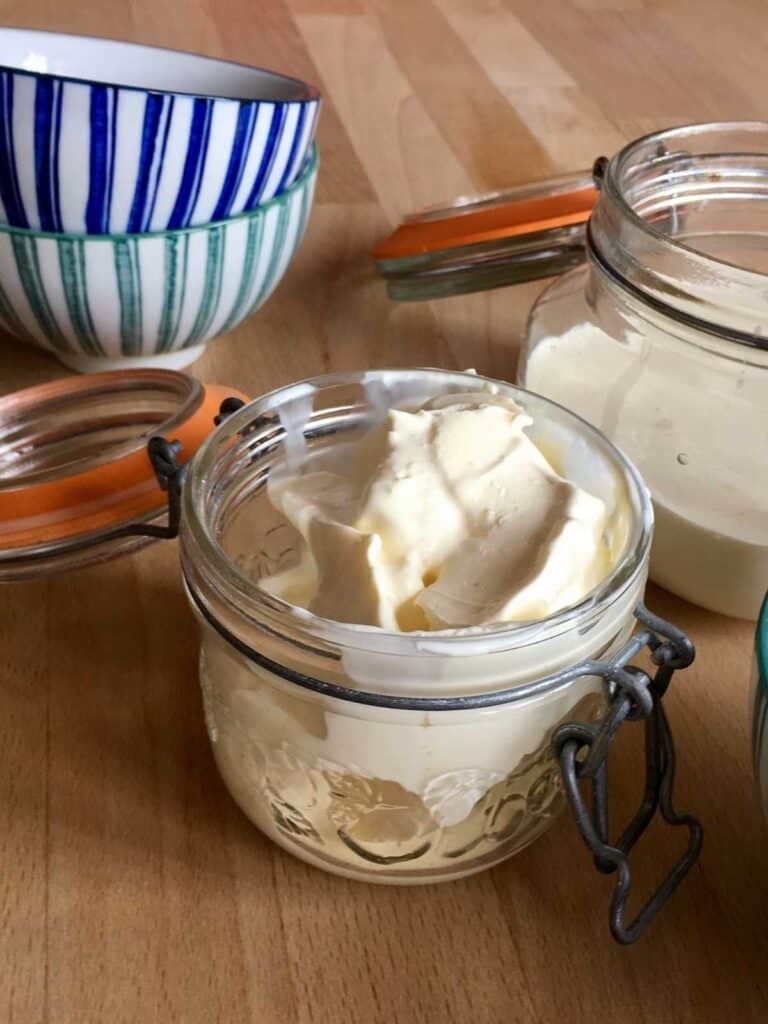 Two glass jars full of clotted cream, the lids are open and there are stripy bowls on the top left of the photo