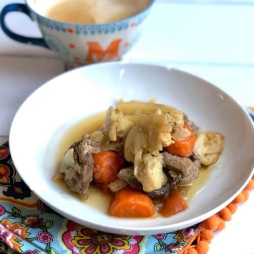 Photo of the pressure cooked pork and cabbage served on a deep white plate with some carrots in the background there is a bright coloured fabric with an orange pompom trim