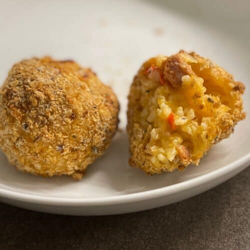 Photo of a couple of Air Fryer Arancini Rice Balls made with chilled left over pressure cooker Chorizo Risotto. The ball on the right has a bite taken out showing the delicious rice inside and the one on the left is whole and showing off a golf-sized crunchy breaded ball of goodness
