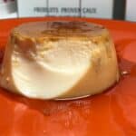 Photo of a pressure cooked flan de Huevo Creme Caramel served on an orange plate, one bite has already been eaten so the smooth inside is showing, there's delicious caramel too