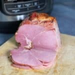 A gammon joint on a wooden chopping board with a Ninja Foodi in the background, three gammon slices rest in front of the joint