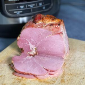 A gammon joint on a wooden chopping board with a Ninja Foodi in the background, three gammon slices rest in front of the joint
