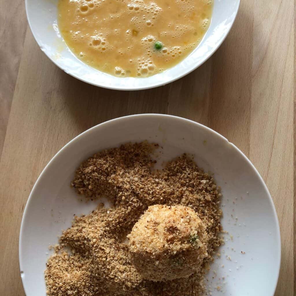 Two white plates from above, the top one has beaten egg and the bottom one has an arancini rice ball coated in breadcrumbs