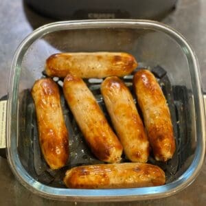Six sausages inside the small glass base of the Ninja CRISPi, they're laid out on the crisper plate, 4 sausages in the middle, 1 at the bottom and 1 at the top