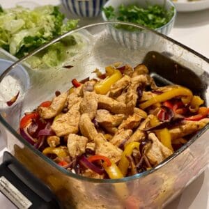 Chicken fajitas inside the large glass base of the Ninja Crispi in the background there are bowls with fresh coriander and lettuce