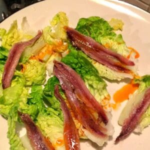 Gem Lettuce leaves draped with anchovies and drizzled with chorizo juices served on a cream color plate