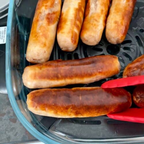 Sausages inside the Ninja Crispi Pro glass dish, with red tongs on the bottom right ready to flip or remove them